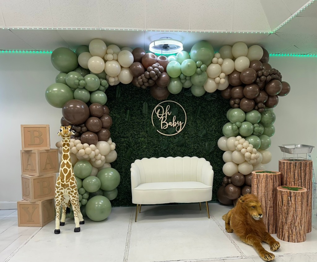 Coming to America themed baby shower backdrop featuring safari decor, balloon arch, lion prop, giraffe prop, and seating area.